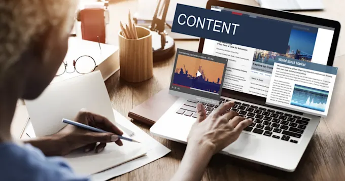 Person writing notes by hand while working on a laptop displaying "CONTENT" webpage graphics with news articles and video thumbnails.