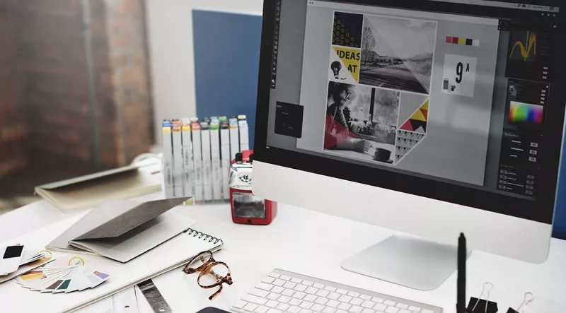 Graphic design workspace with a computer monitor displaying a layout project, surrounded by markers, color swatches, notebooks, and glasses on a desk.