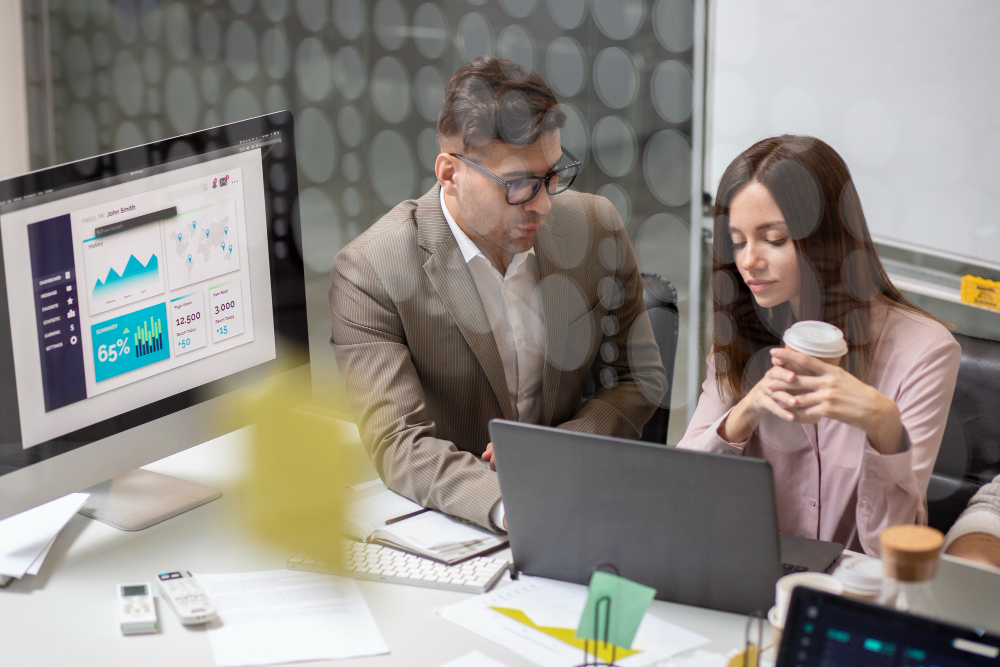 Colleagues reviewing a laptop together in an office, with a monitor displaying a business analytics dashboard in the foreground.