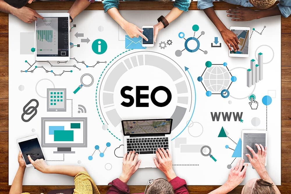 Overhead view of a group using laptops, tablets, and smartphones around a table with an "SEO" concept graphic featuring web, analytics, and network icons.